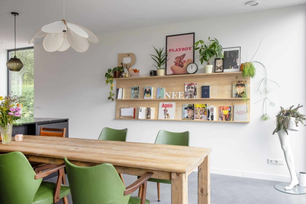 Eetgedeelte met een houten tafel en groene stoelen in Zijdewind. Achter de tafel hangt een kastje met daarin posters, platen, planten en andere accessoires. Er is gekozen voor een gevlinderde betonvloer.