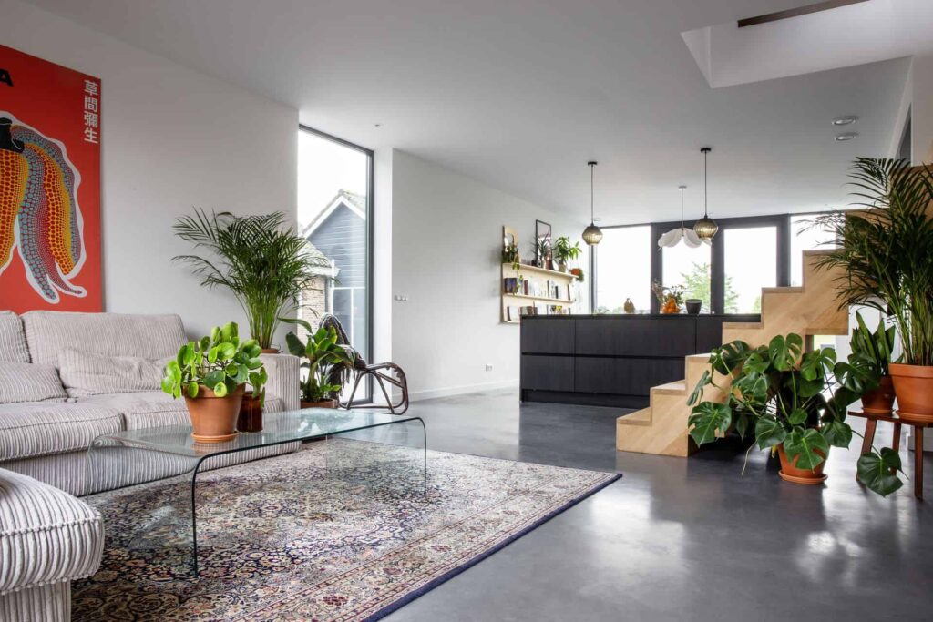 Kijkje in de woning in Zijdewind met een gevlinderde betonvloer. Vooraan zie je de roze bank, een gekleurd vloerkleed en wat planten. Daarachter staat een houten trap en de zwarte keuken.