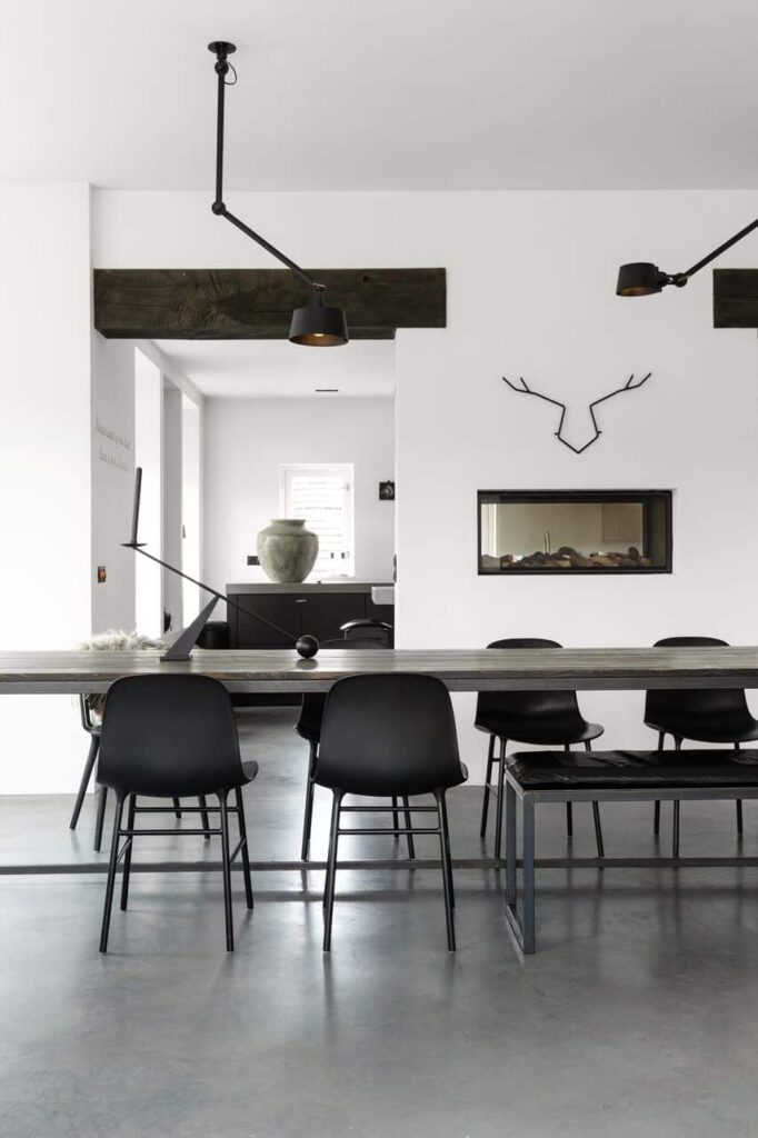 Houten tafel met een stalen frame, gecombineerd met zwarte stoelen en een gevlinderde betonvloer. De wanden zijn wit en in 1 wand zit een haard met hierboven een zwart metalen hert.