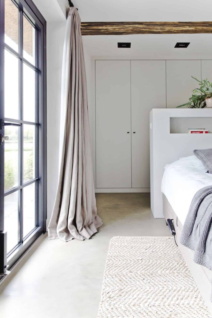 Slaapkamer met een gevlinderde betonvloer met daarop een beige bed, een licht vloerkleed en lichte gordijnen.