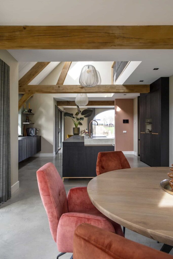 Nieuw gebouwde boerderij in Horst met een gevlinderde betonvloer. In combinatie met een zwarte keuken, houten balken aan het plafond, een houten tafel met daarom roze stoelen.